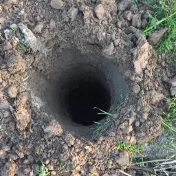 Okrągły otwór w ziemi otoczony grudami ziemi i zieloną trawą, widoczny fragment buta w dolnym rogu.