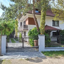 Dwupiętrowy dom z brązowym dachem i białymi ścianami, częściowo zasłonięty przez drzewo z pnączami, widoczny zza ozdobnego, kutego ogrodzenia z kostki brukowej.