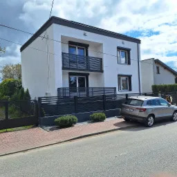 Nowoczesny, dwukondygnacyjny dom z białą elewacją i grafitowymi akcentami, widoczny z poziomu ulicy obok zaparkowanego samochodu.