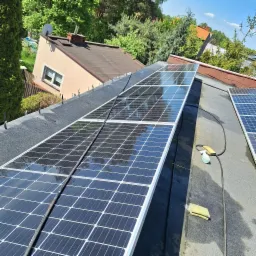 Panele słoneczne na płaskim dachu z widocznymi narzędziami czyszczącymi i odbiciem drzew w panelach.