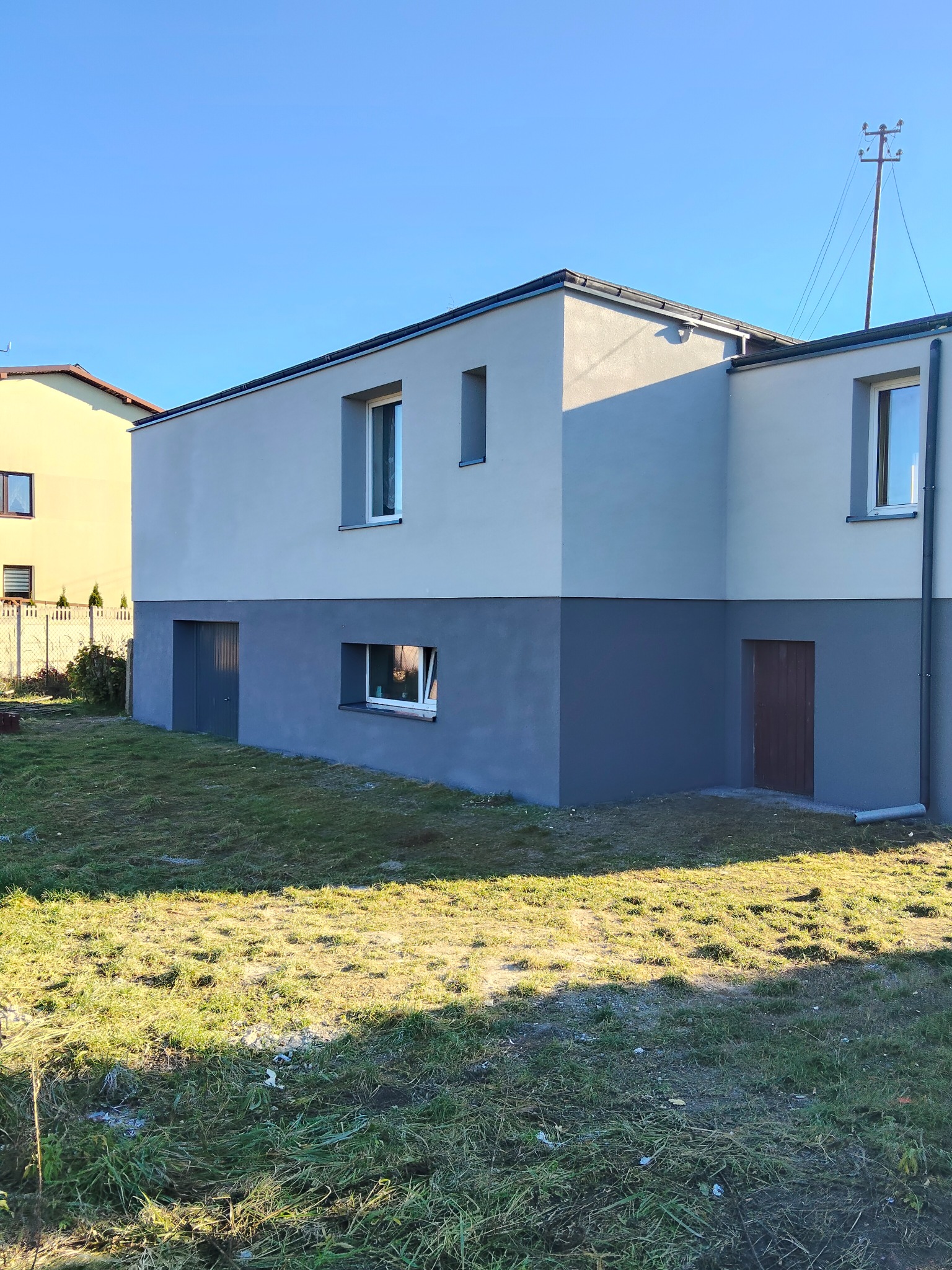 Dwukolorowa elewacja domu z szarą dolną częścią i jaśniejszą górną, widok z trawnika w słoneczny dzień. Widoczne okna i drzwi.