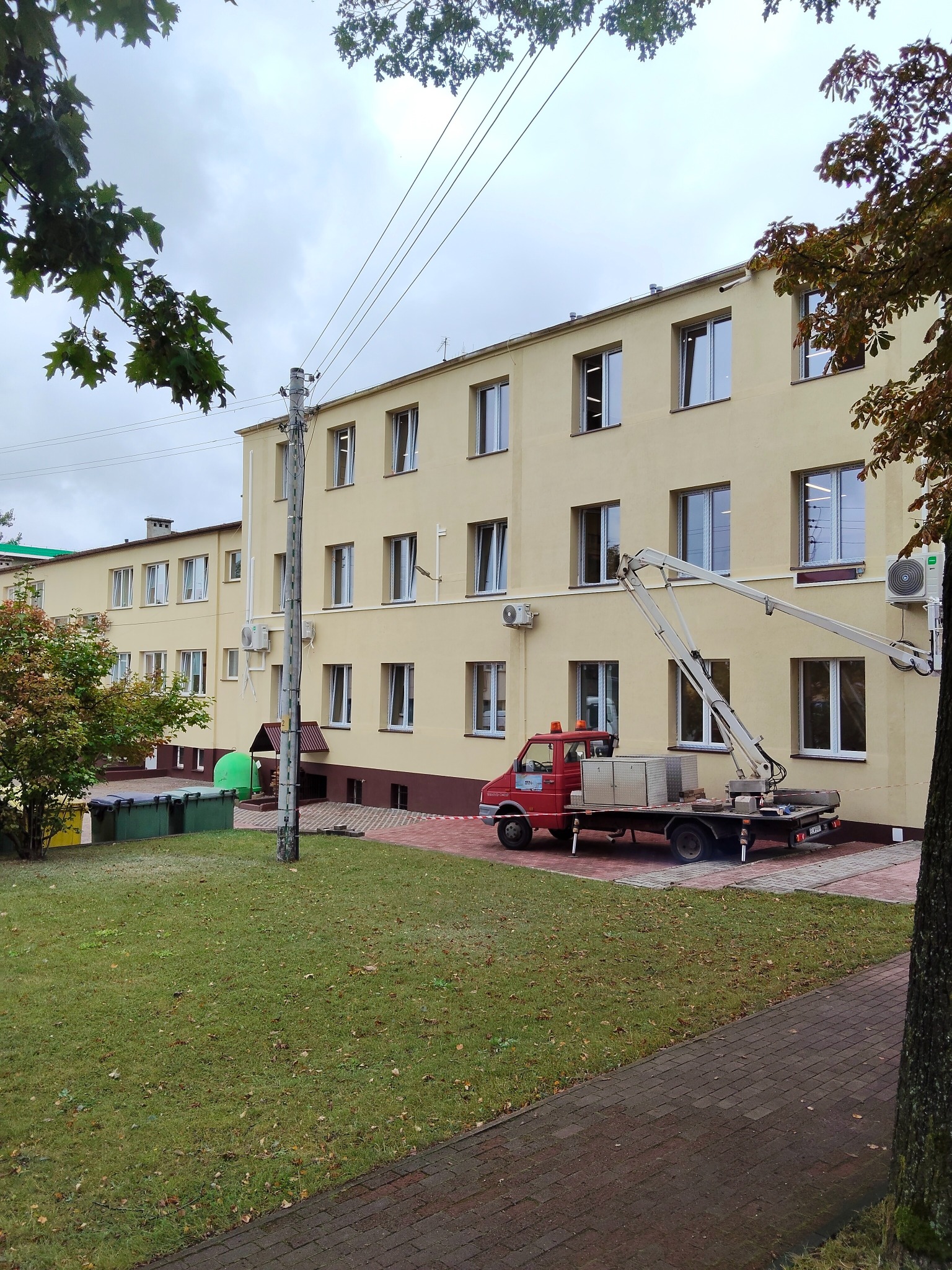 Żółty budynek z czerwonym samochodem z podnośnikiem. Prace na wysokościach przy klimatyzatorze. Widok z poziomu chodnika, pochmurne niebo.