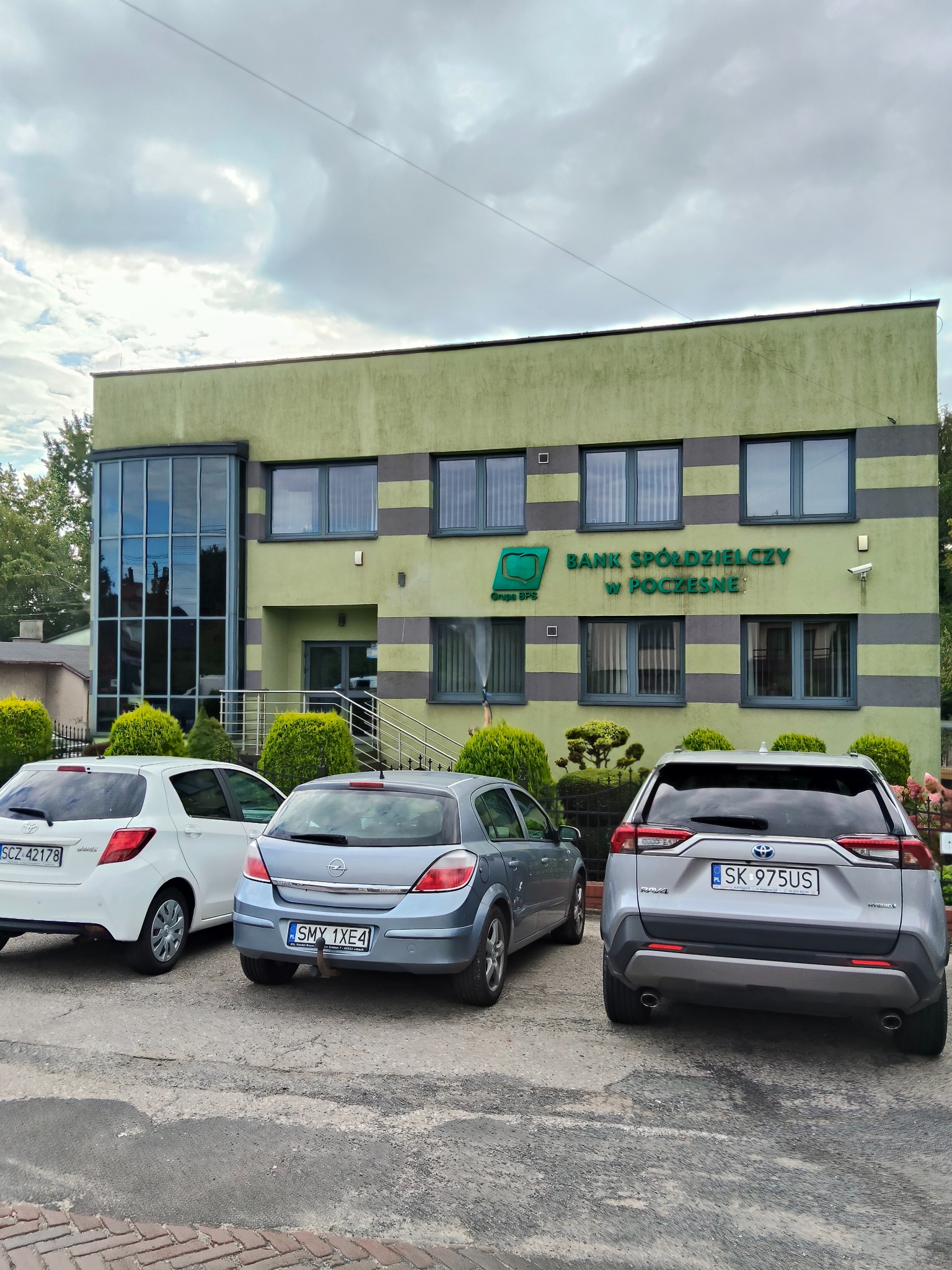 Budynek Banku Spółdzielczego w Poczesnej z zielono-szarymi pasami, widoczny z parkingu z trzema samochodami. Uwagę zwraca nowoczesna szklana fasada po lewej stronie.