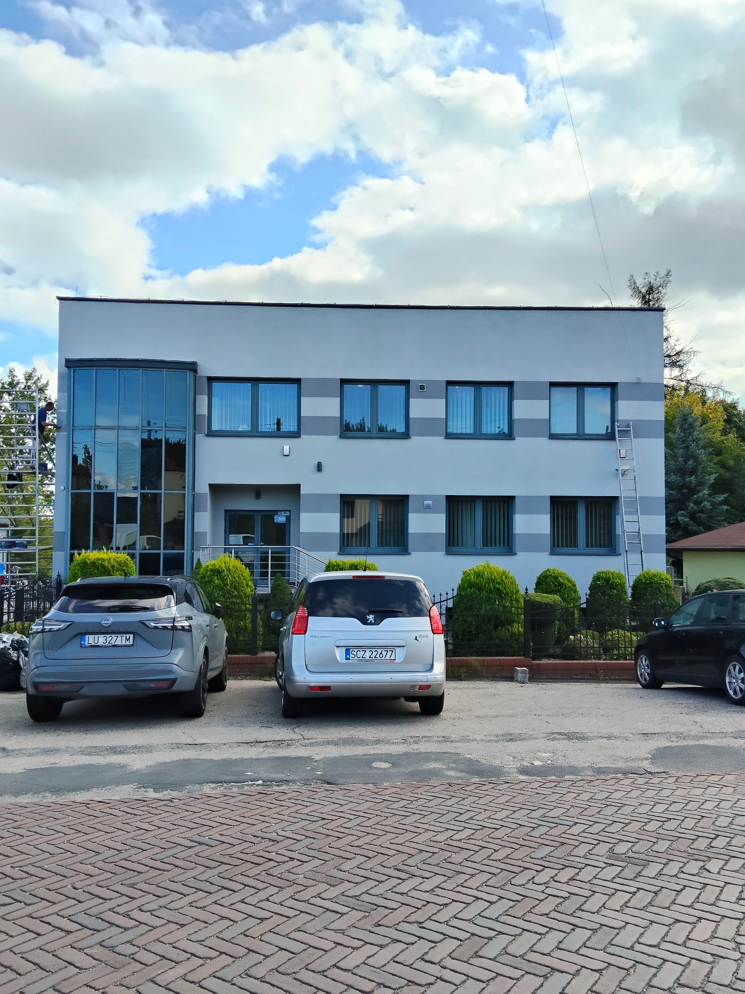 Dwupiętrowy budynek biurowy z szarą elewacją i ciemnymi oknami. Na elewacji widoczne poziome pasy. Przed budynkiem zaparkowane samochody i zadbana zieleń.