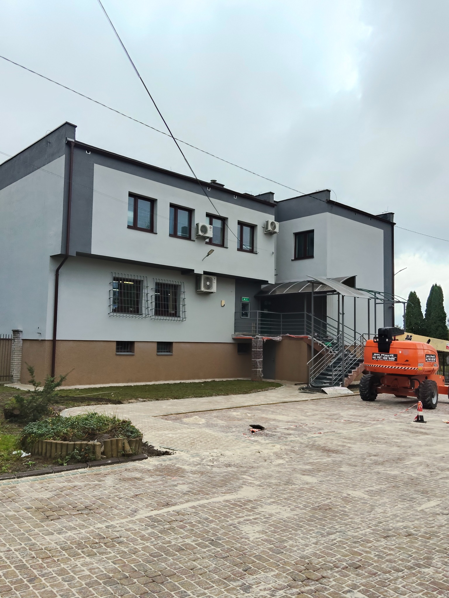 Dwukondygnacyjny budynek po renowacji elewacji w odcieniach szarości, z pomarańczowym podnośnikiem koszowym na kostce brukowej. Widoczne klimatyzatory i zieleń.