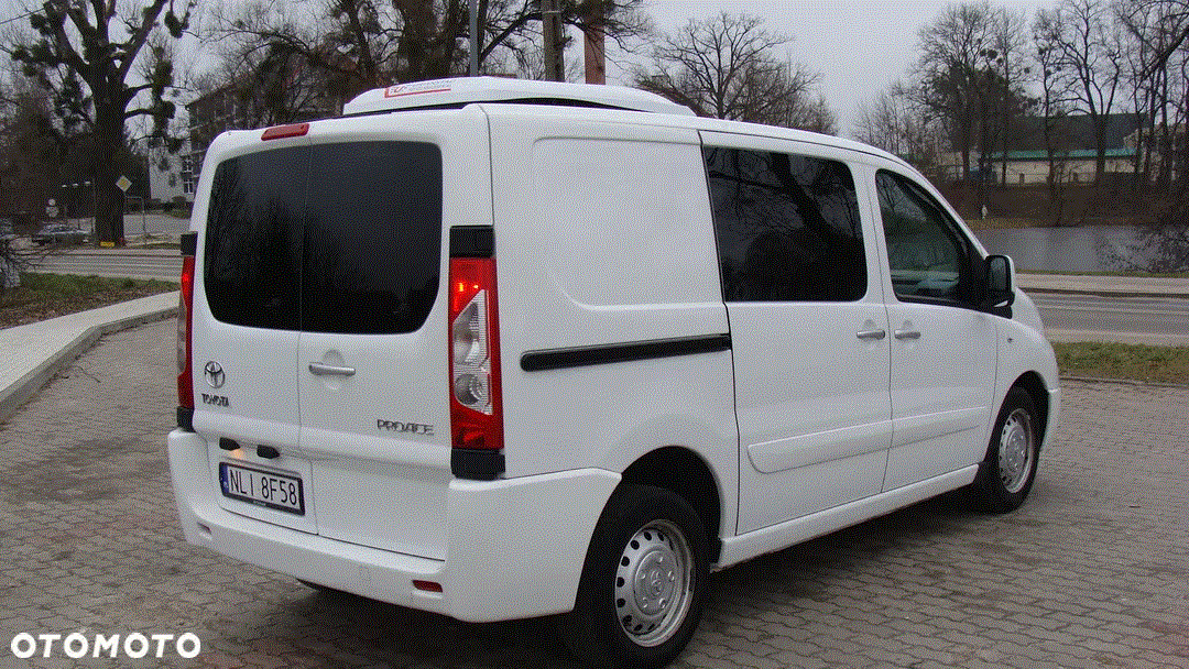 Biały bus Toyota ProAce z chłodnią na dachu, widok z tyłu, zaparkowany na brukowanym placu przy drodze.