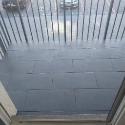 Szare płytki gresowe ułożone na balkonie, widok z góry przez metalową balustradę na parking z samochodami.