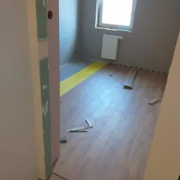 Pokój w trakcie układania paneli podłogowych, widoczne klipsy dystansowe, częściowo ułożona podłoga z paneli drewnopodobnych, fragment ściany z szarą farbą i okno z grzejnikiem.