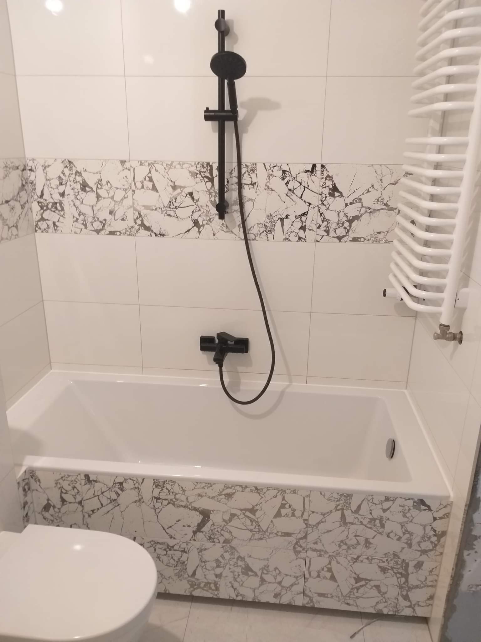 Biała wanna obudowana płytkami imitującymi marmur, czarna bateria prysznicowa i grzejnik drabinkowy w wykończonej łazience z białymi płytkami.