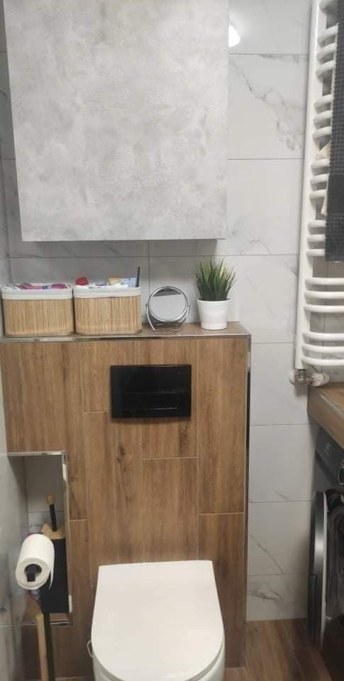 Nowoczesna toaleta w zabudowie z drewnopodobnych paneli, biała ceramika sanitarna, szare płytki imitujące marmur na ścianach, rattanowe koszyki na blacie, dekoracyjna roślina w doniczce, grzejnik...