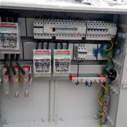 Otwarta biała skrzynka elektryczna z zainstalowanymi bezpiecznikami, rozłącznikami i przewodami, widoczne elementy montażowe i przyłączeniowe.