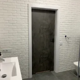 Ciemne drzwi wewnętrzne z białą ościeżnicą w łazience z białą cegłą na ścianach i szarą podłogą, widoczna umywalka i toaleta.