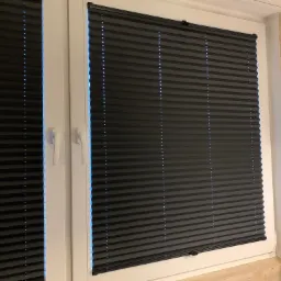 Zasłonięte okno z białymi ramami i ciemnymi plisowanymi żaluzjami w pomieszczeniu z panelami ściennymi imitującymi drewno.