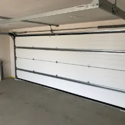 Nowa, biała segmentowa brama garażowa z widocznymi prowadnicami i sprężynami naciągowymi, zamontowana w surowym garażu z betonową podłogą i ż&oacute;łtą taśmą malarską przy ścianach.