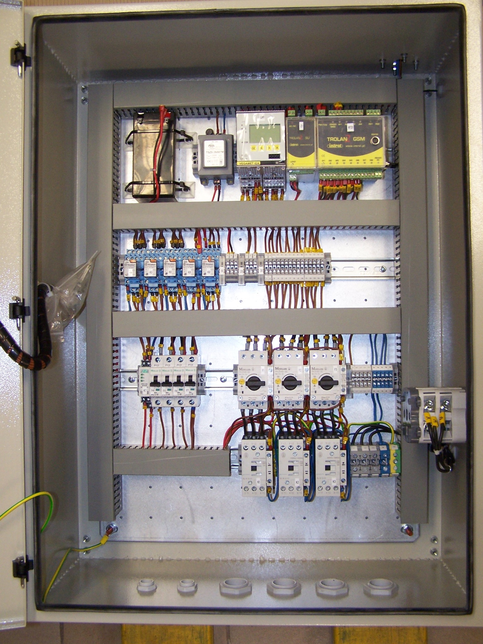 Otwarta szafa sterownicza z widocznymi elementami elektrycznymi, okablowaniem i zabezpieczeniami, zainstalowanymi na szynach DIN, z żółto-zielonym przewodem uziemiającym w lewym dolnym rogu.