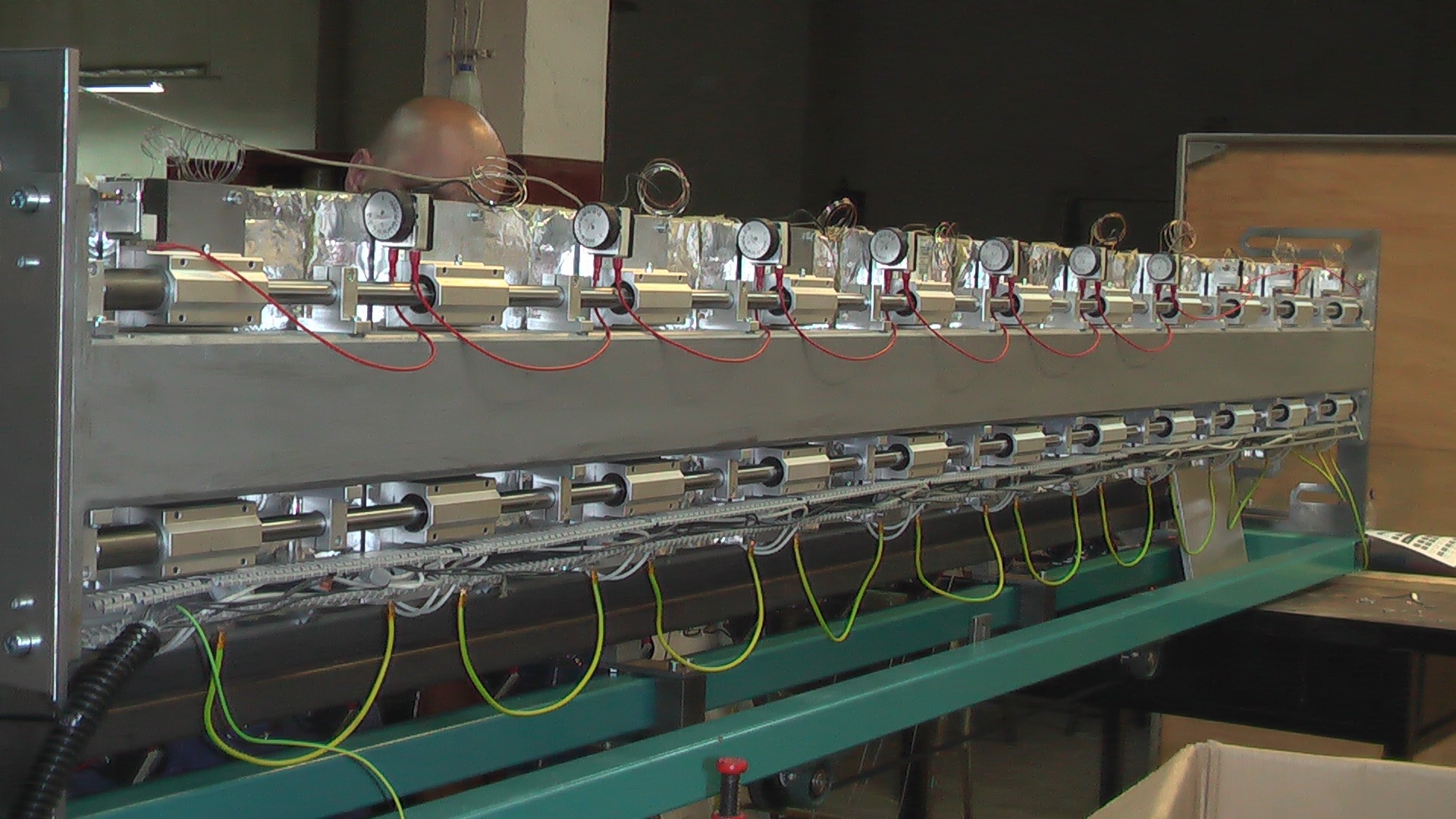 Szczegółowe ujęcie fragmentu maszyny przemysłowej z rzędem cylindrów pneumatycznych, przewodami i manometrami, widok z boku z fragmentem głowy majsterkowicza w tle.