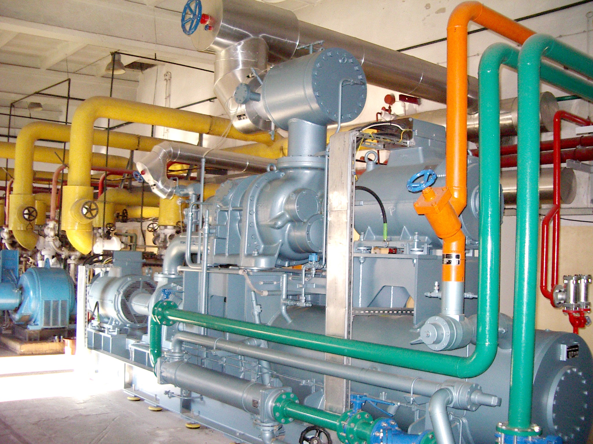 Kompresor przemysłowy z systemem rur w kolorach żółtym, zielonym i pomarańczowym, widoczne zawory z niebieskimi pokrętłami, oświetlenie sufitowe.