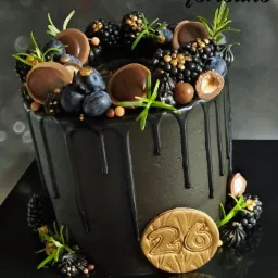 Czarny tort z polewą i dekoracją z jeżyn, borówek, czekoladek i rozmarynu, ozdobiony złotym medalionem z numerem 26.