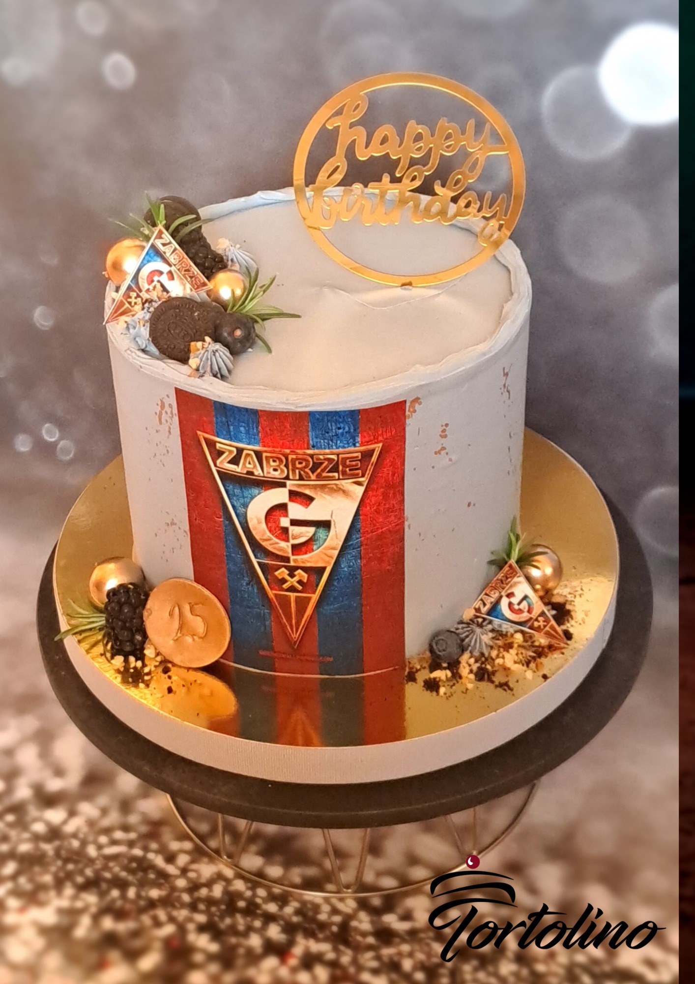 Tort urodzinowy z motywem klubu sportowego Górnik Zabrze, dekorowany złotym napisem 'Happy Birthday', kulkami, ciasteczkami i flagą klubu, umieszczony na złotej paterze.