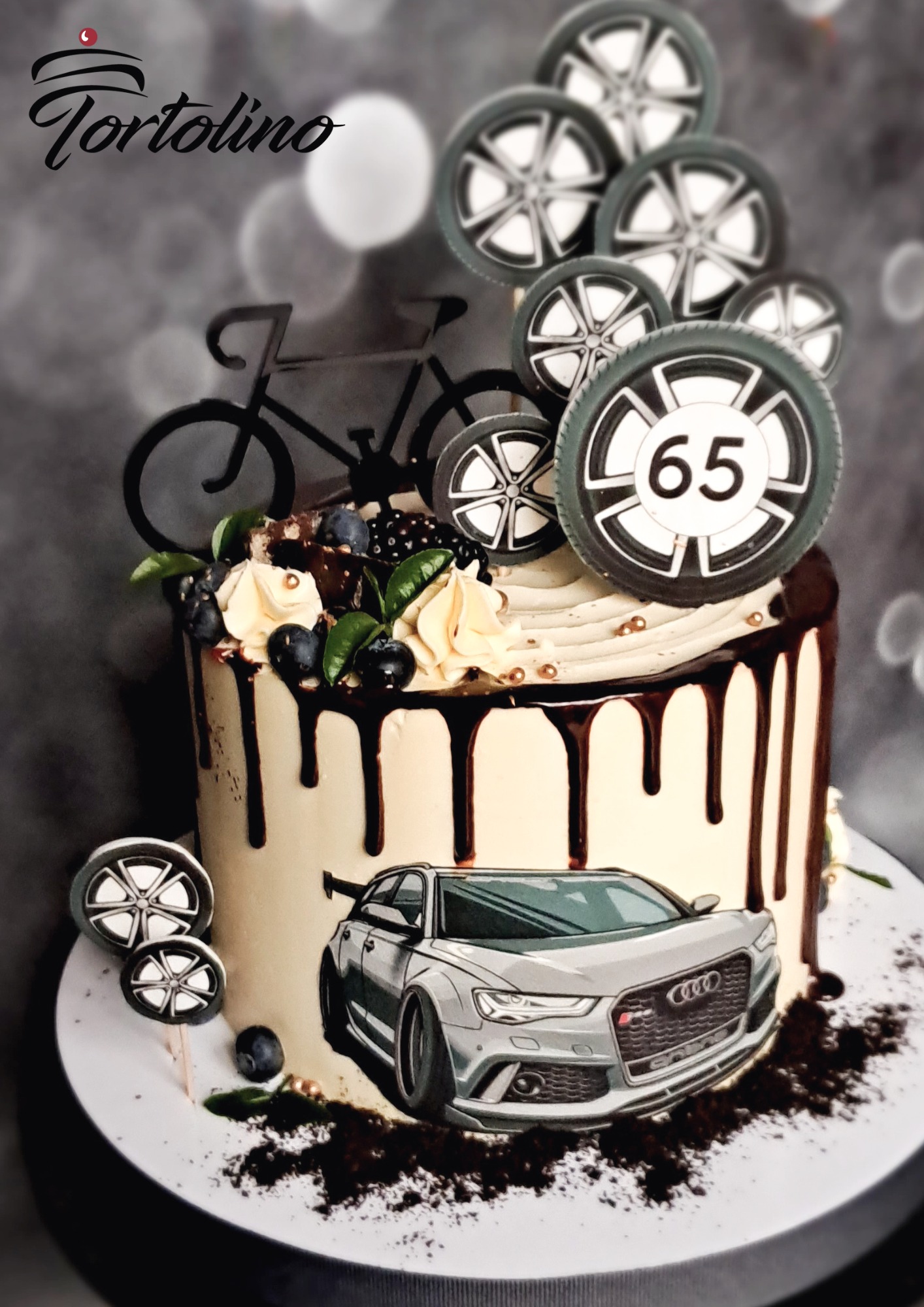 Tort zdobiony motywami motoryzacyjnymi: autko, koła z numerem 65, rower, oblany czekoladą, z kremowymi rozetkami i owocami leśnymi, na rozmytym tle.
