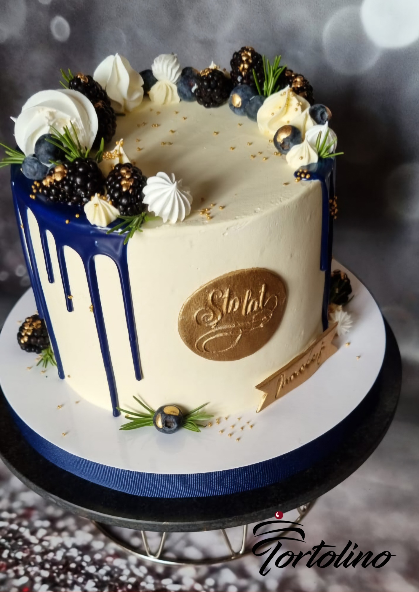 Tort na stulecie z granatową polewą, jeżynami, borówkami, bezikami i rozmarynem, umieszczony na paterze z granatową wstążką.