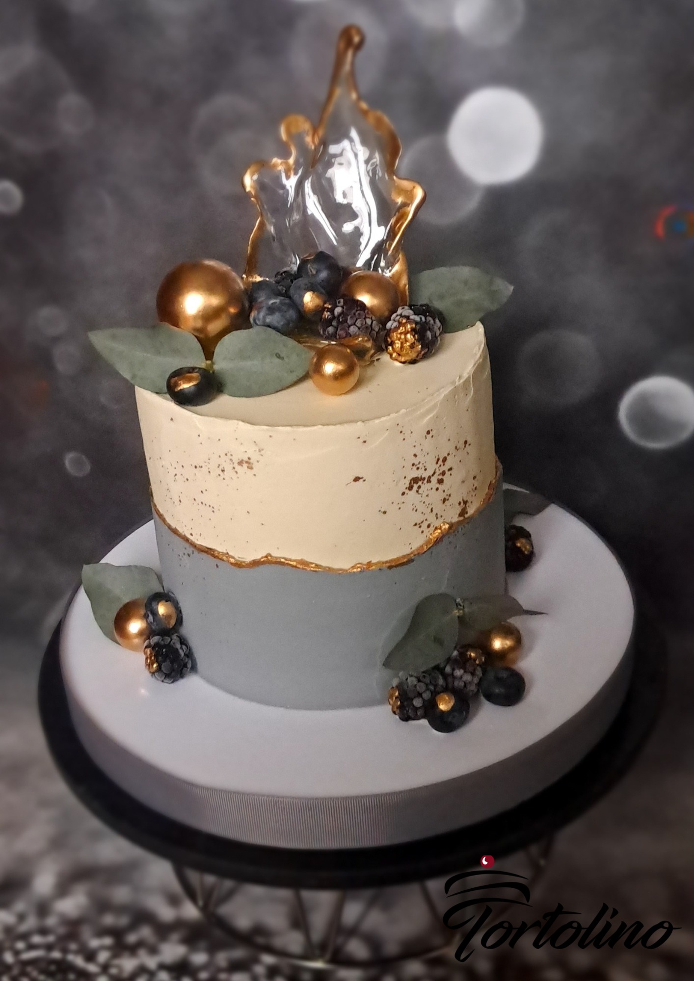 Dwupoziomowy tort w odcieniach szarości i kremu, zdobiony złotymi kulkami, jeżynami, borówkami i liśćmi eukaliptusa, z przezroczystą, karmelową dekoracją na wierzchu.