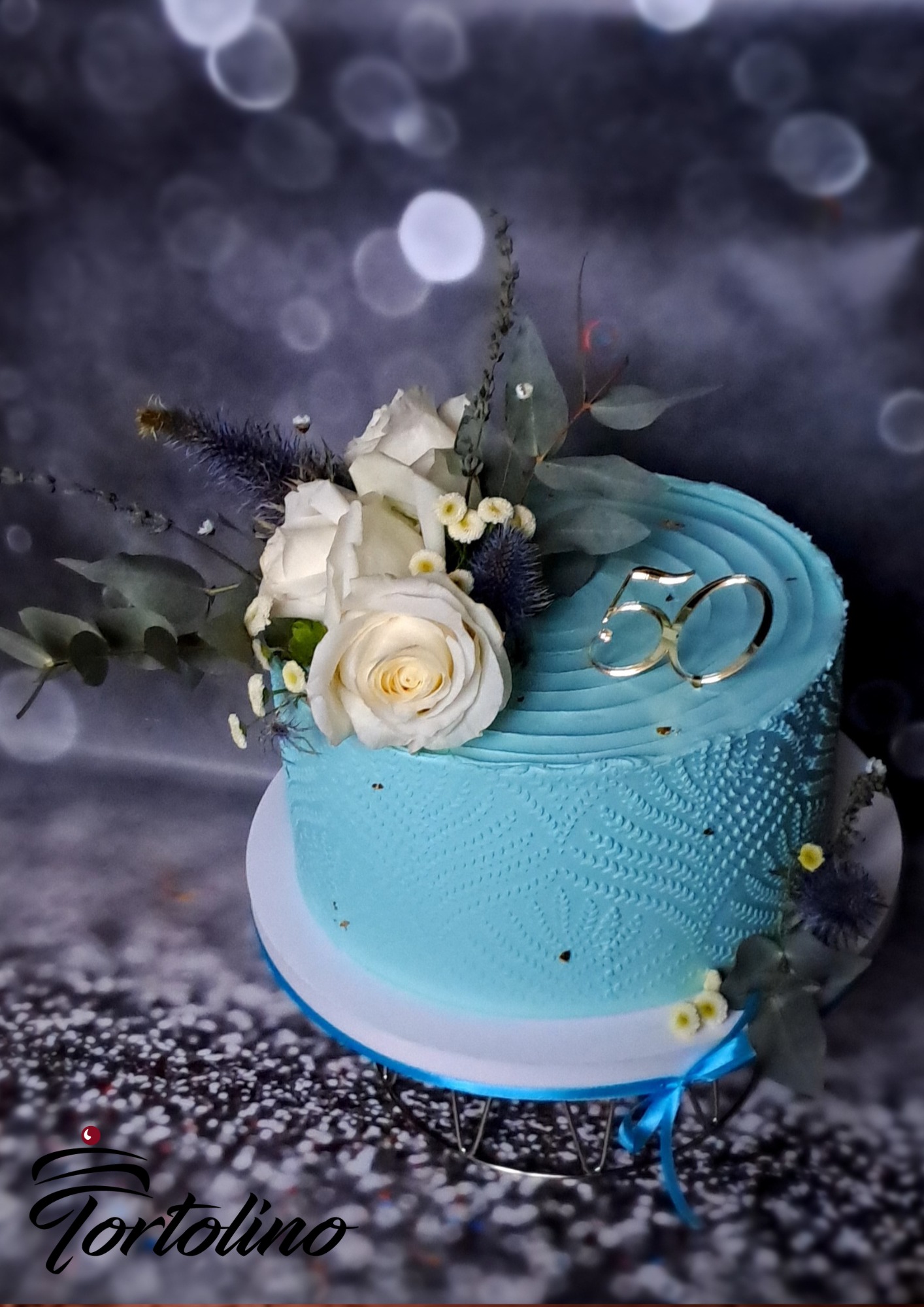 Niebieski tort urodzinowy na 50-te urodziny, udekorowany białymi różami i złotym napisem '50', umieszczony na białej paterze z niebieską wstążką.