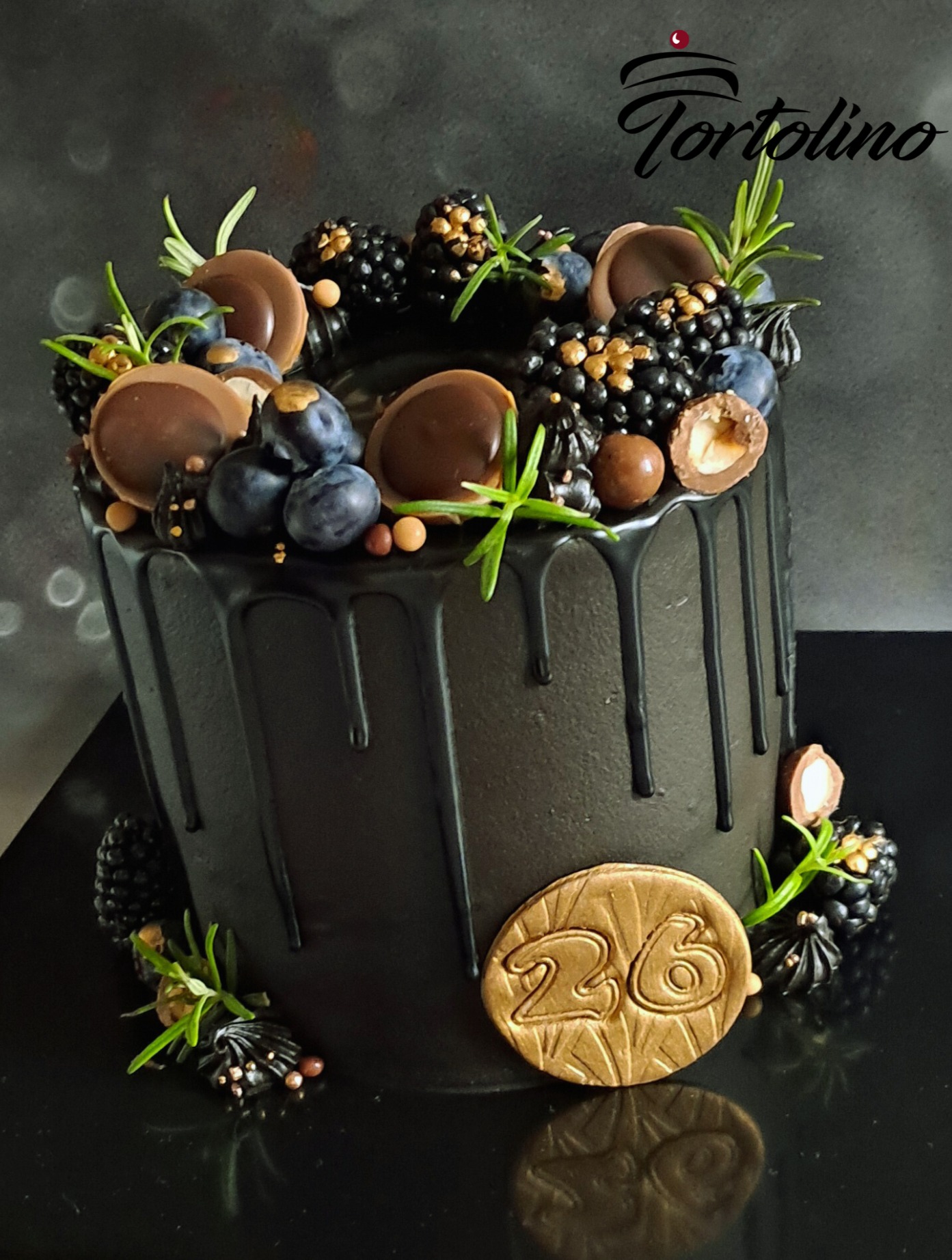 Elegancki tort urodzinowy w czarnej polewie z dripami, udekorowany świeżymi jeżynami, borówkami, czekoladowymi dyskami i rozmarynem, z okrągłą dekoracją z numerem 26 w kolorze złota.
