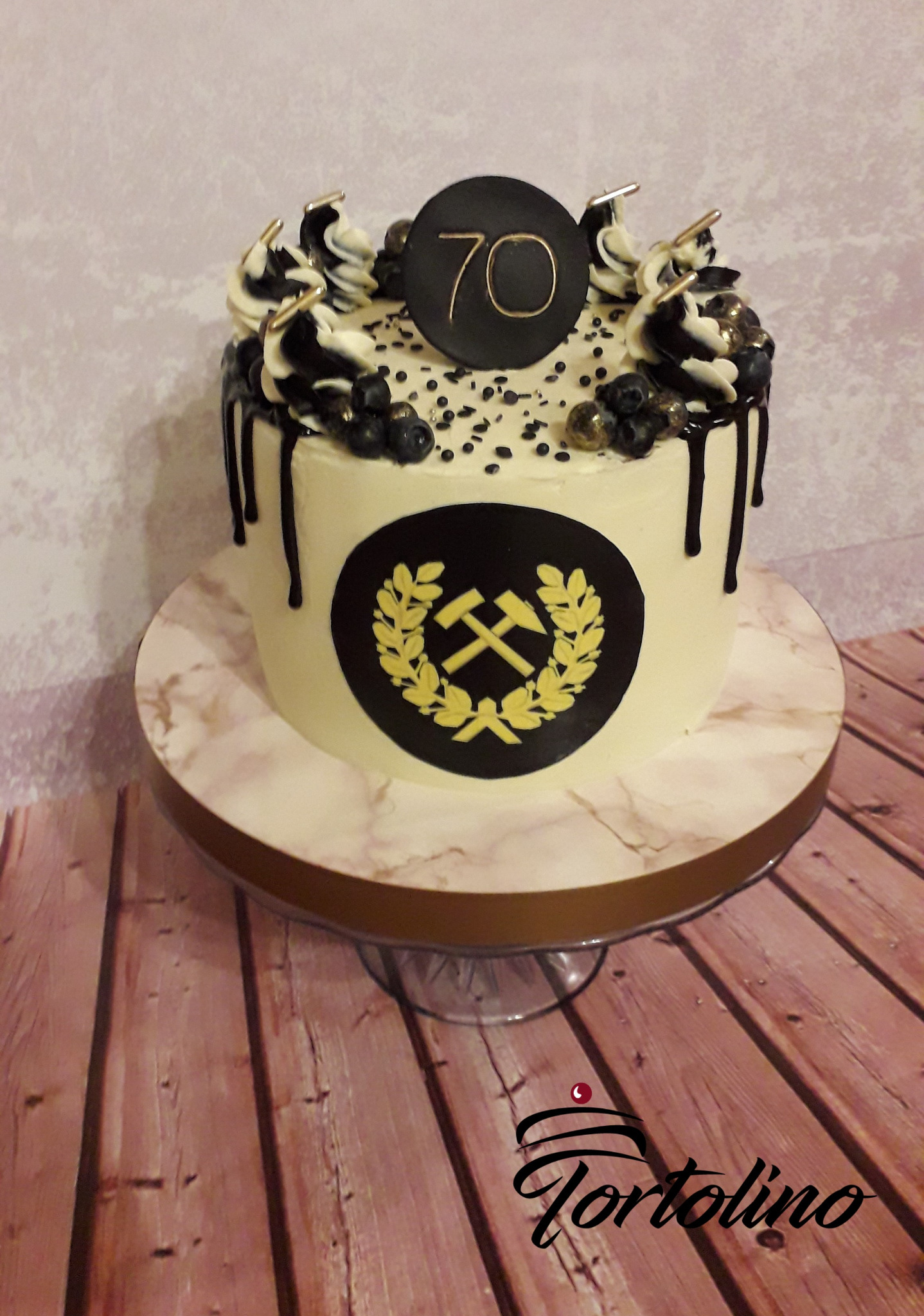 Tort na 70. urodziny z symbolem górniczym, dekorowany czarnym lukrem, borówkami i złotymi akcentami, umieszczony na marmurowym stojaku.
