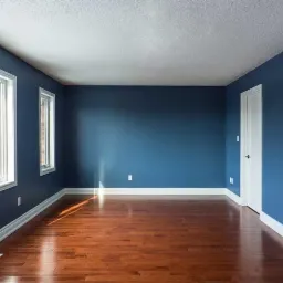 Wnętrze pokoju po remoncie: ściany pomalowane na niebiesko, drewniana podłoga, białe listwy przypodłogowe, okno z widokiem na budynek z czerwonej cegły, uchwyt ścienny na telewizor.