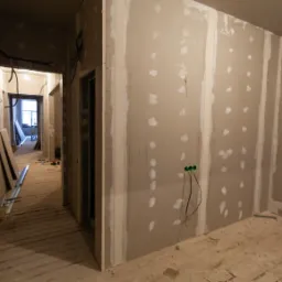 Wnętrze w trakcie prac z płytami kartonowo-gipsowymi, widoczne szpachlowane łączenia, wystające przewody elektryczne z gniazdek oraz drewniana podłoga.