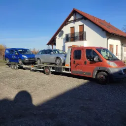 Pomarańczowy samochód laweta z dwoma samochodami osobowymi na platformie, zaparkowany na żwirowym podwórku przed białym domem z czerwoną dachówką w słoneczny dzień.