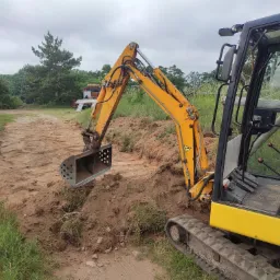 Żółta mini koparka z łyżką zagłębia się w ziemię, przygotowując teren pod budowę, widoczny świeży wykop i częściowo zarośnięte otoczenie.