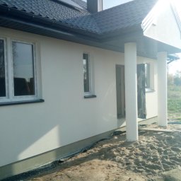 TermoDom Usługi Marcin Oracki - Świeżo otynkowana elewacja domu jednorodzinnego z białym tynkiem, widoczne okna z białymi ramami, ciemna dachówka i kolumny podtrzymujące zadaszenie wejścia, teren wokół domu częściowo zasypany...