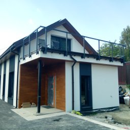 TermoDom Usługi Marcin Oracki - Nowoczesny dom z elewacją w kolorach bieli i antracytu, wykończony drewnopodobną okładziną, z balkonem na dachu; prace wykończeniowe w toku, widoczne materiały budowlane i pracownik porządkujący...