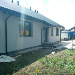 TermoDom Usługi Marcin Oracki - Świeżo otynkowana elewacja domu jednorodzinnego z widocznymi śladami prac wykończeniowych na trawniku, w tle zaparkowane samochody dostawcze.