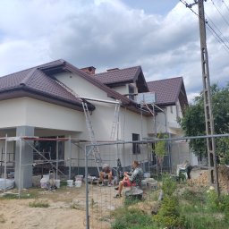 TermoDom Usługi Marcin Oracki - Dwóch pracowników w przerwie siedzi na tle domu w trakcie prac elewacyjnych, widoczne rusztowania, drabiny i wiaderka z farbą, elewacja w jasnym odcieniu szarości, brązowy dach.