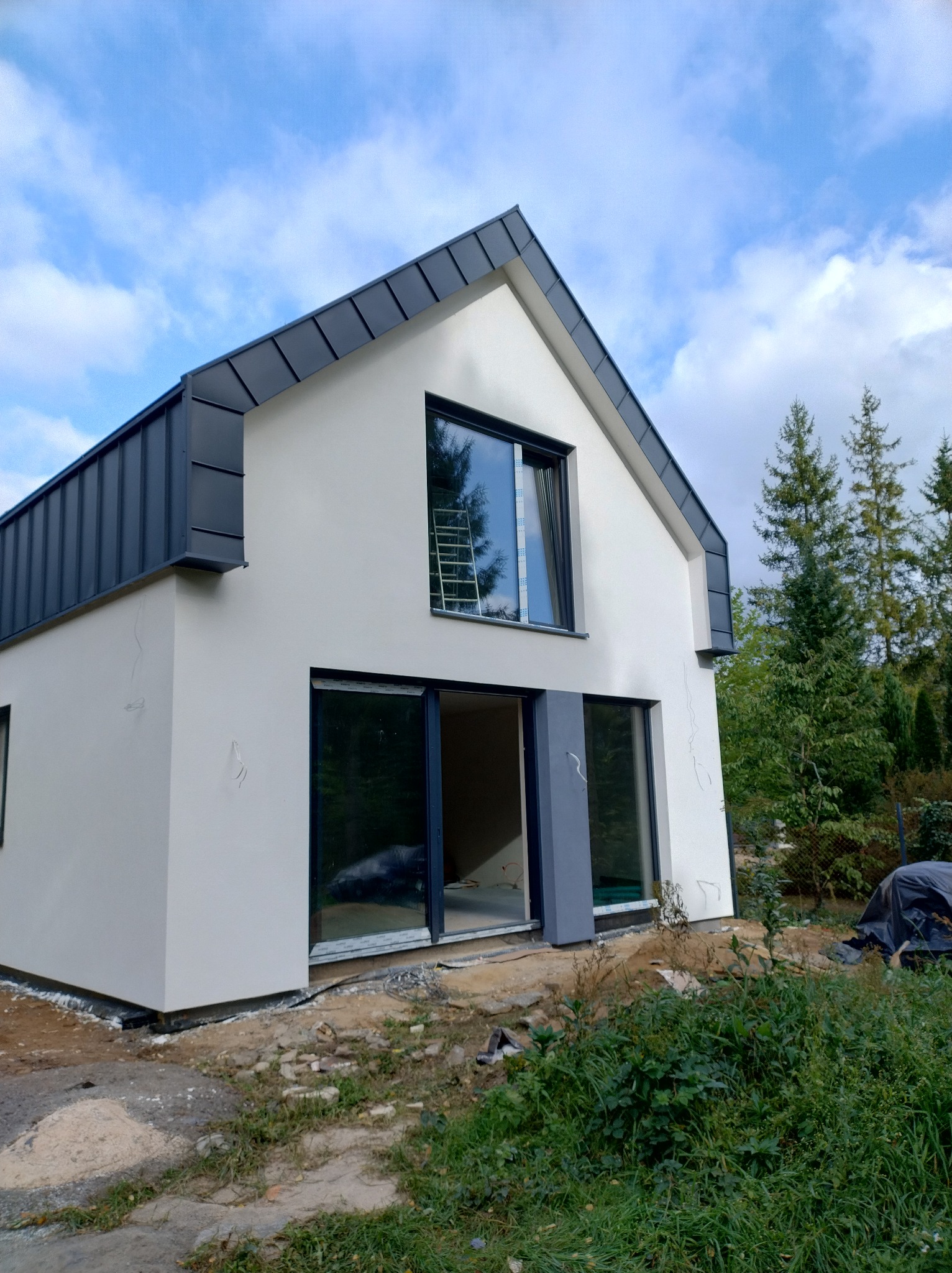 Nowoczesny dom jednorodzinny z białą elewacją i grafitowym dachem, widoczne pęknięcia na ścianie, teren wokół domu w trakcie prac wykończeniowych.