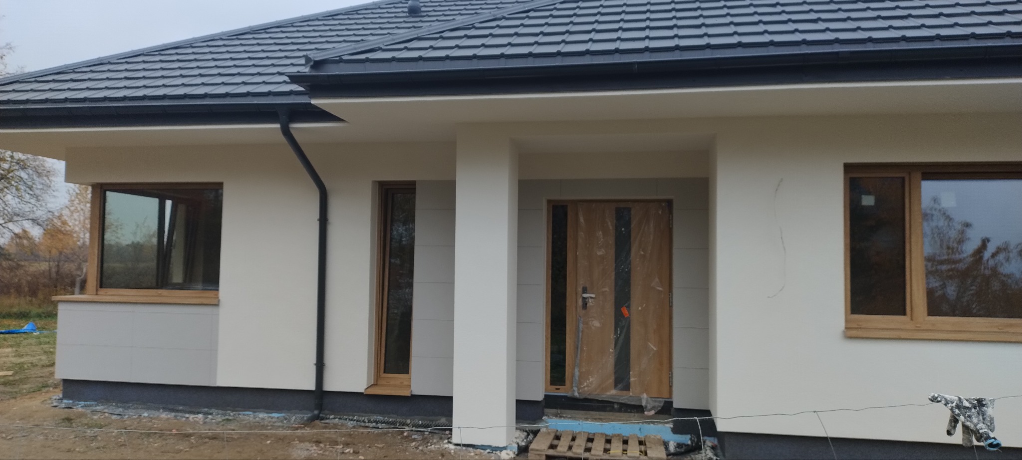 Nowoczesny dom jednorodzinny z jasną elewacją, drewnianymi oknami i grafitowym dachem. Wejście osłonięte kolumnami, drzwi zabezpieczone folią. Widoczne detale wykończenia.