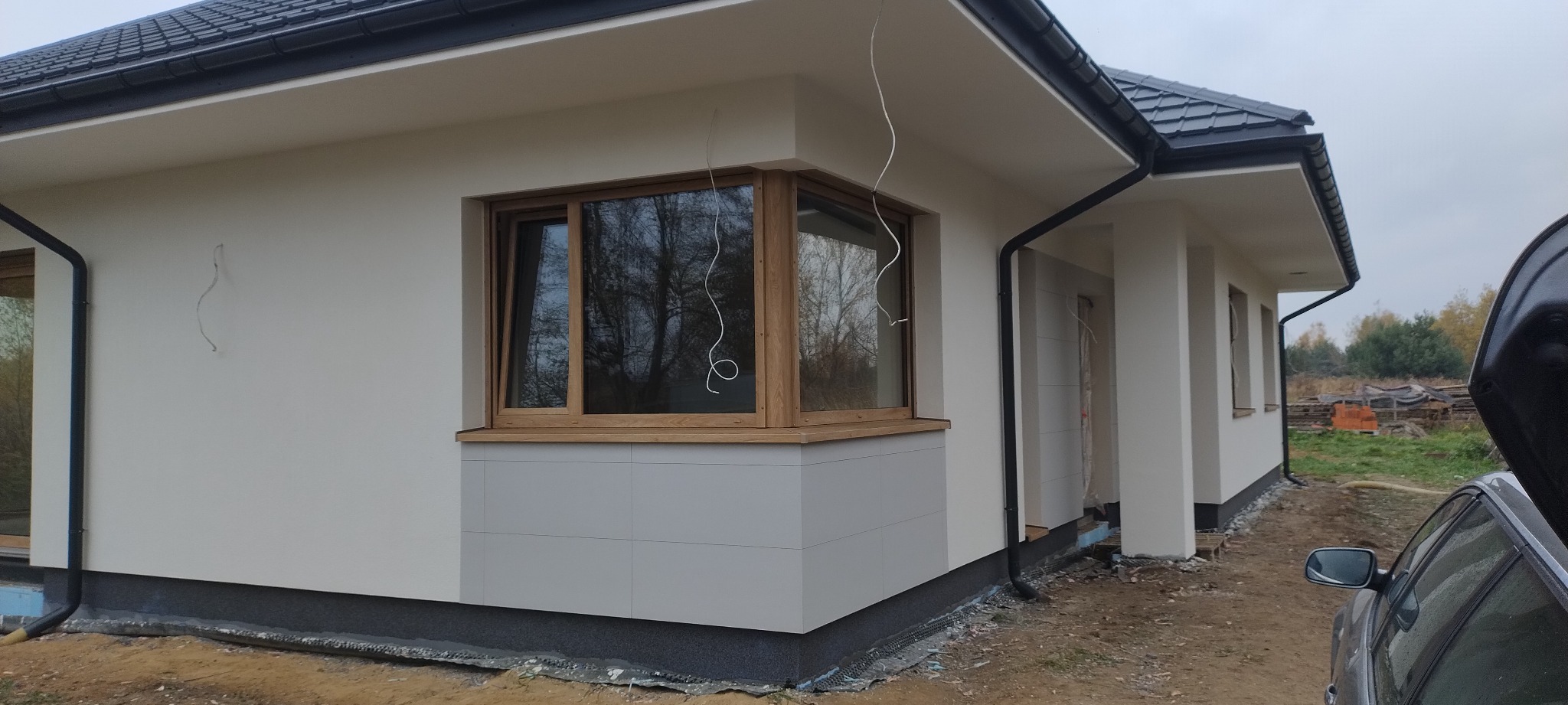 Nowoczesny dom jednorodzinny z oknem narożnym i fragmentem elewacji wykończonym płytami w odcieniach szarości. Widoczne elementy orynnowania i teren wokół budynku.