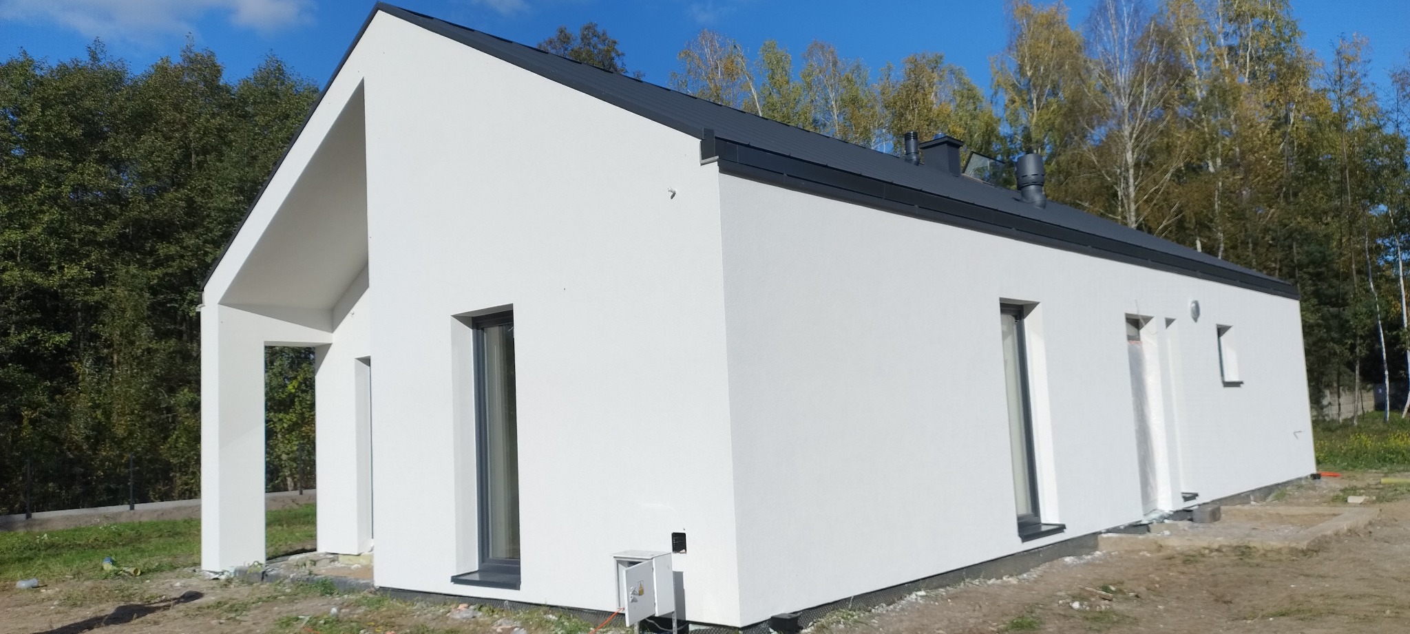 Biała, nowoczesna elewacja domu jednorodzinnego z ciemnym dachem na tle zieleni. Widoczne okna i elementy wykończeniowe. Minimalistyczny styl.