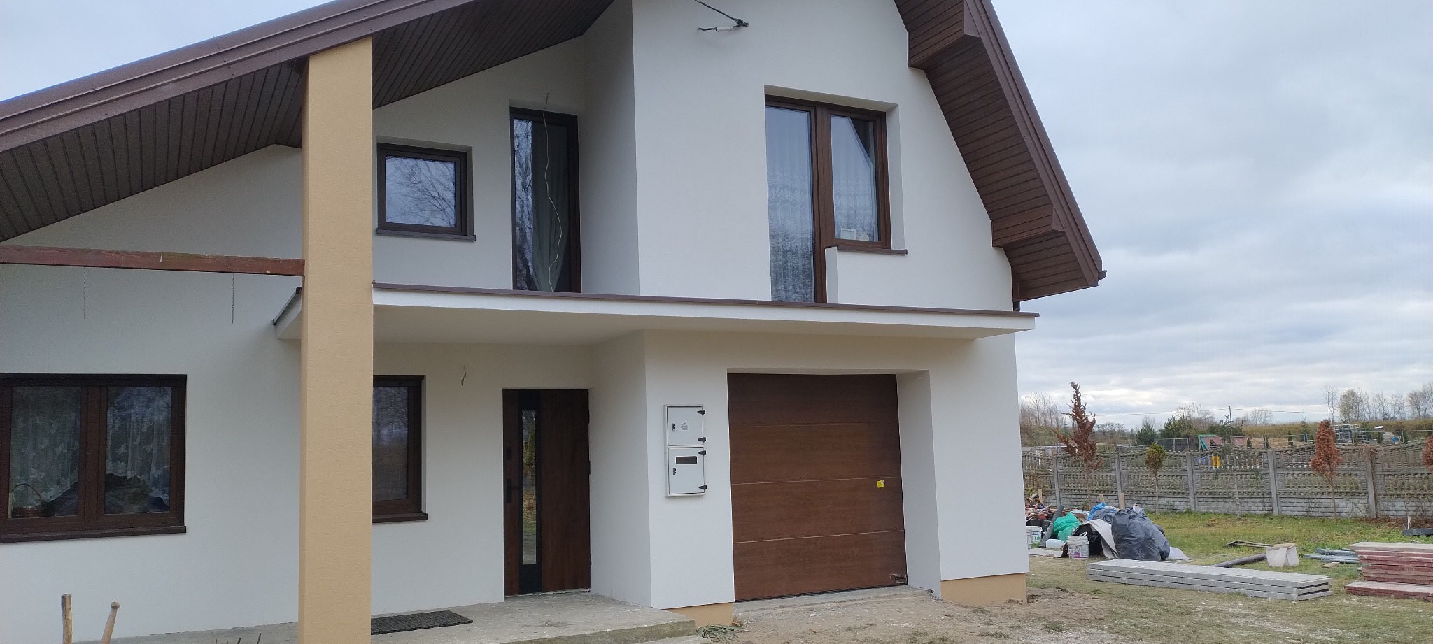 Dwukondygnacyjny dom z jasną elewacją, brązowymi oknami i bramą garażową. Widoczne elementy wykończeniowe i otoczenie budynku. Ujęcie z poziomu gruntu.