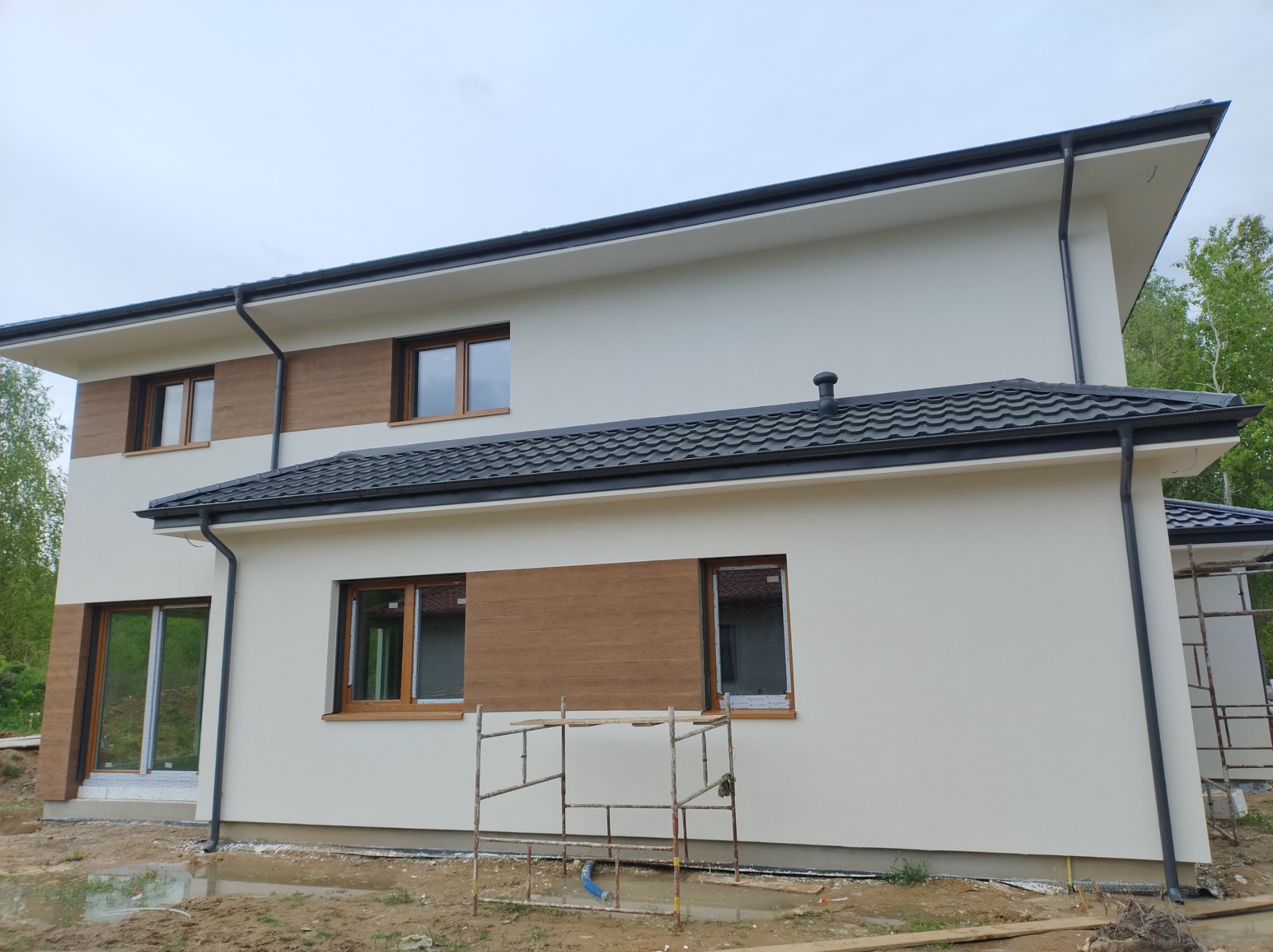 Dwukondygnacyjny dom z jasną elewacją, drewnianymi panelami dekoracyjnymi, ciemną dachówką i rynnami, stojący na placu budowy z widocznymi rusztowaniami i kałużami.
