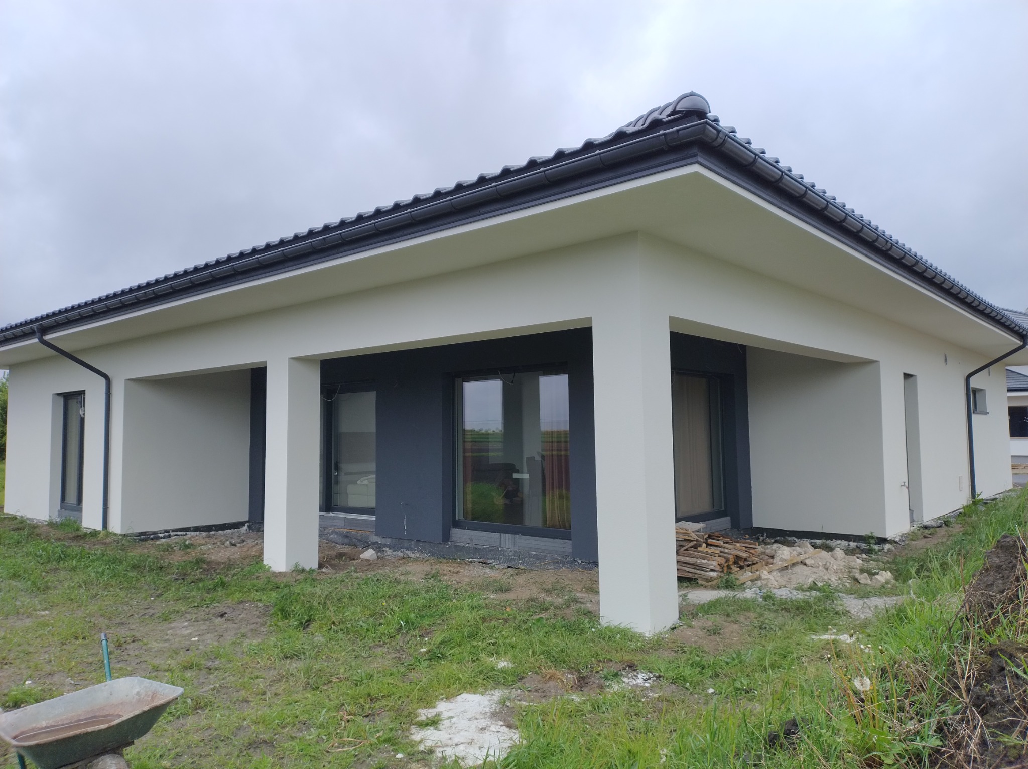 Wykończona elewacja domu jednorodzinnego w odcieniach bieli i szarości, z widocznymi elementami krajobrazu przed budynkiem i stertą drewna w pobliżu.