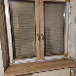 F.H.U.Michał Tomera - Zamknięte, dwuskrzydłowe okno z profili w odcieniu dębu Turner, z zamontowanym parapetem MDF w tym samym kolorze. Widoczne rolety wewnętrzne i surowe wykończenie ściany.