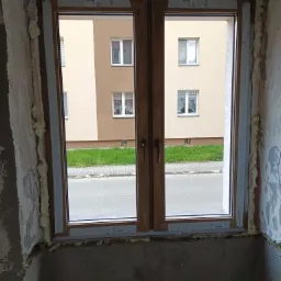 Zamontowane, dwuskrzydłowe okno z drewnopodobnymi ramami, widok na budynek z zielenią. Pianka montażowa wypełnia szczeliny. Surowe ściany w tle.