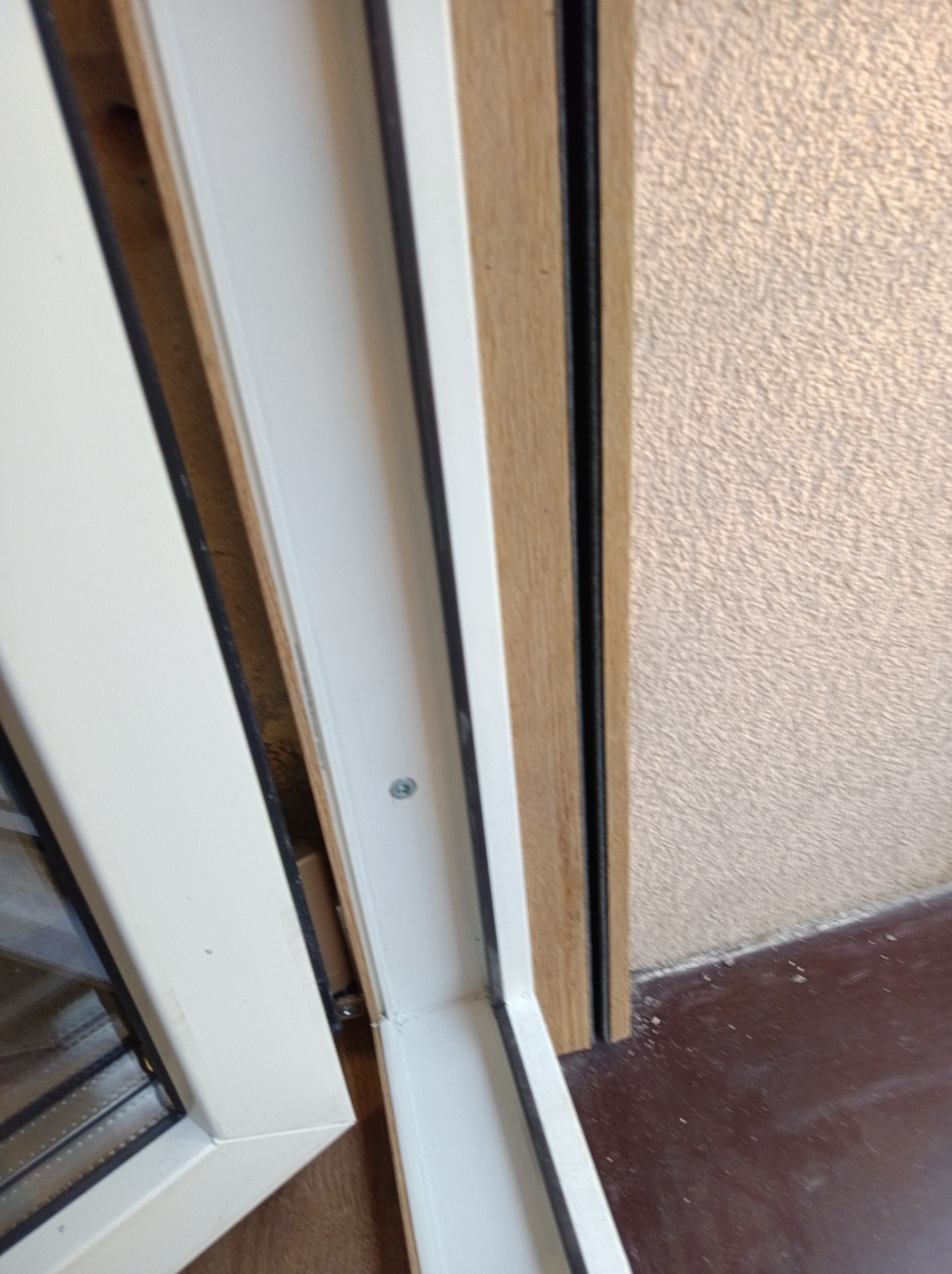 Detal montażu białego okna z drewnianą ramą, widoczny fragment ściany o fakturze baranka, śruba mocująca i brązowa podłoga. Ujęcie z bliska, z góry.