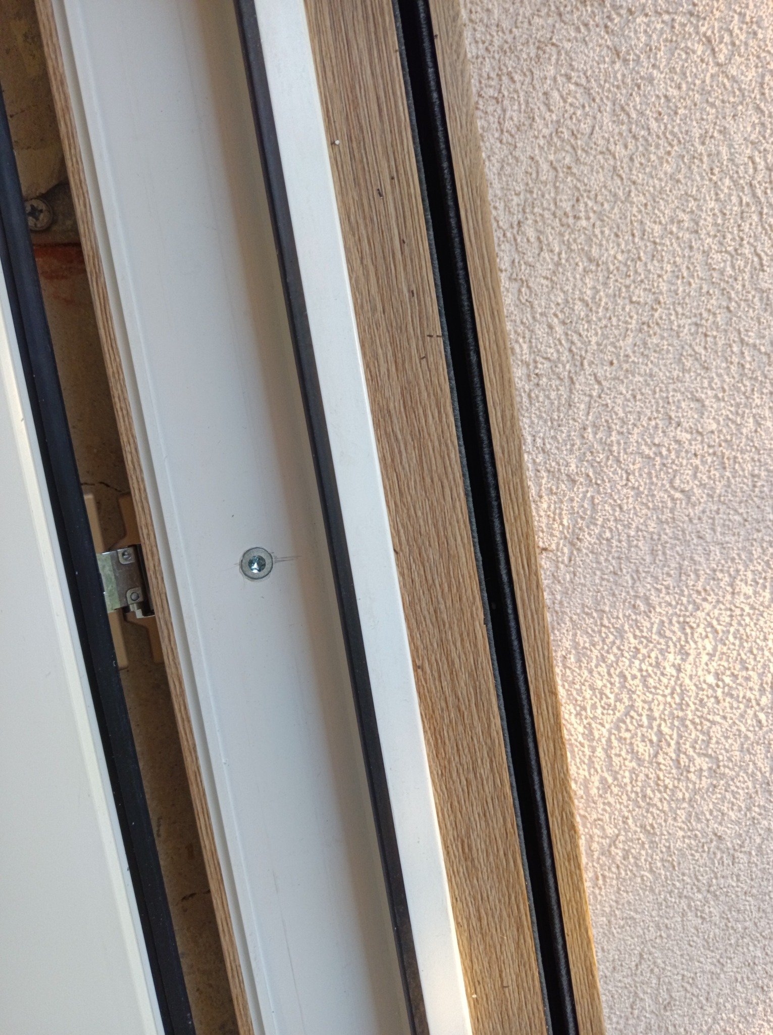 Fragment okna PCV w kolorze białym z drewnopodobną okleiną, widoczny zamek i uszczelka, na tle chropowatej ściany. Detal montażu okna.