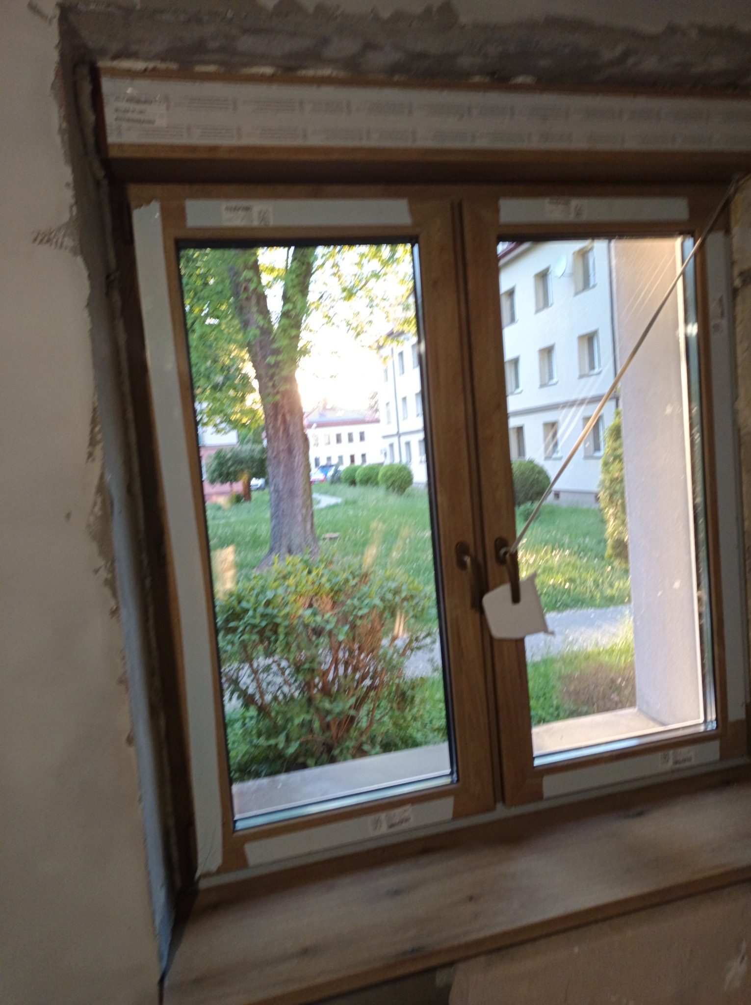 Montaż dwuskrzydłowego okna z drewnianą ramą. Widok na zieleń i budynek przez szybę. Świeżo osadzony parapet, surowy mur wokół okna.