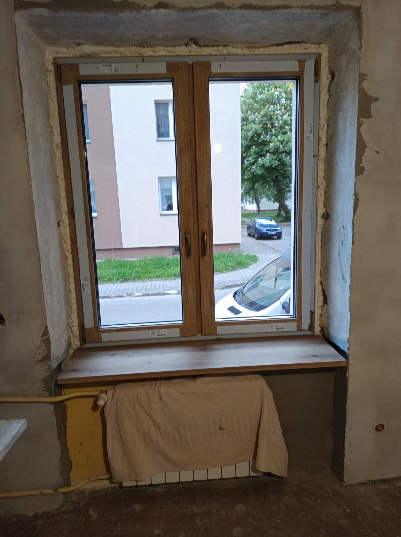 Montaż okna z drewnianą ramą w surowym wnętrzu. Widoczne pianka montażowa, drewniany parapet i fragment grzejnika z zasłoną. Na zewnątrz budynek i zaparkowane auto.
